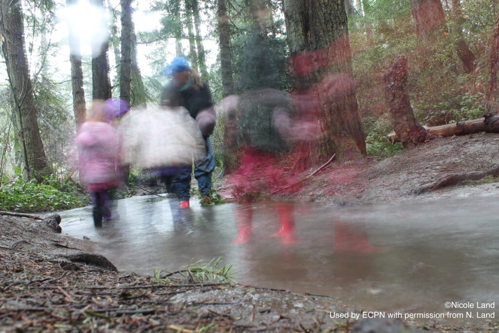 Several people, including children, are blurred, walking through a forested area with a shallow stream.