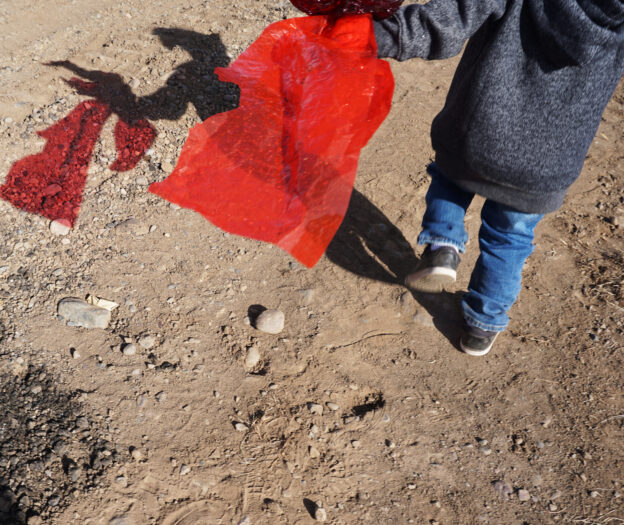 Child walking on dirt path, holding a red translucent fabric casting a vivid shadow on the ground.